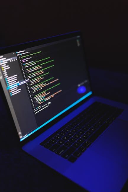 A person with short dark hair, wearing a light blue shirt, is seated at a white desk working on a silver laptop that displays a screen filled with lines of colorful code in shades of blue, red, yellow