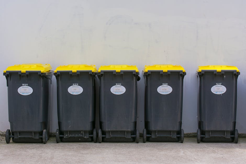 An outdoor scene showing a large pile of mixed waste and rubbish spilling onto a paved area in front of a commercial building. The waste includes cardboard boxes, plastic bags, paper, and packaging ma